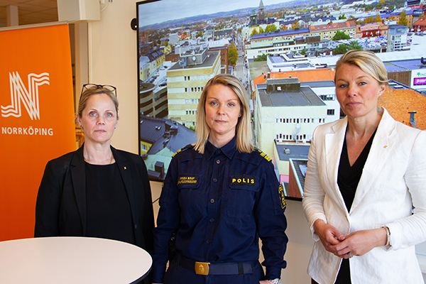 Tre kvinnor, som allvarligt ser in i kameran, står framför vepa med Norrköpings kommuns logga. Kvinnan i mitten har polisuniform på sig.