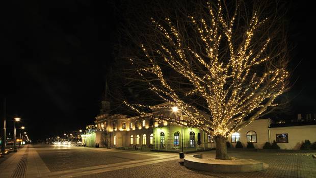 Ett träd, smyckat med ljusslingor, står framför ett stationshus. 
