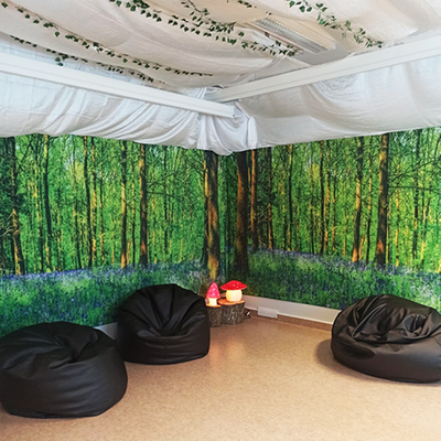 Ett rum med fototapet som föreställer en skog. På golvet finns sittpuffar. I taket hänger vitt tyg och girlanger med gröna blad.